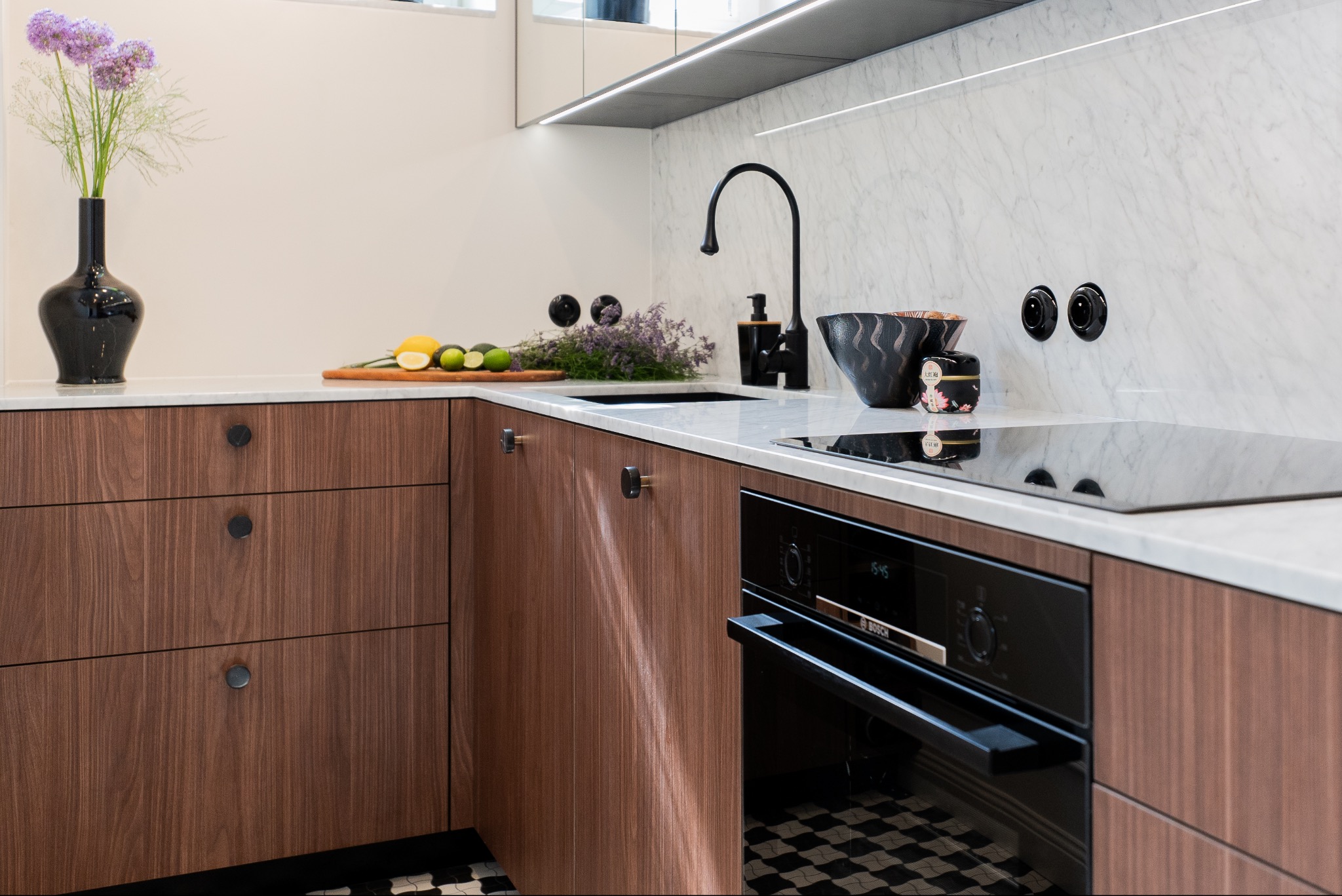 Nowoczesna kuchnia z drewnianymi szafkami, marmurowym blatem, czarną armaturą i czarno-białą podłogą w szachownicę; na blacie wazon z fioletowymi kwiatami i owoce.