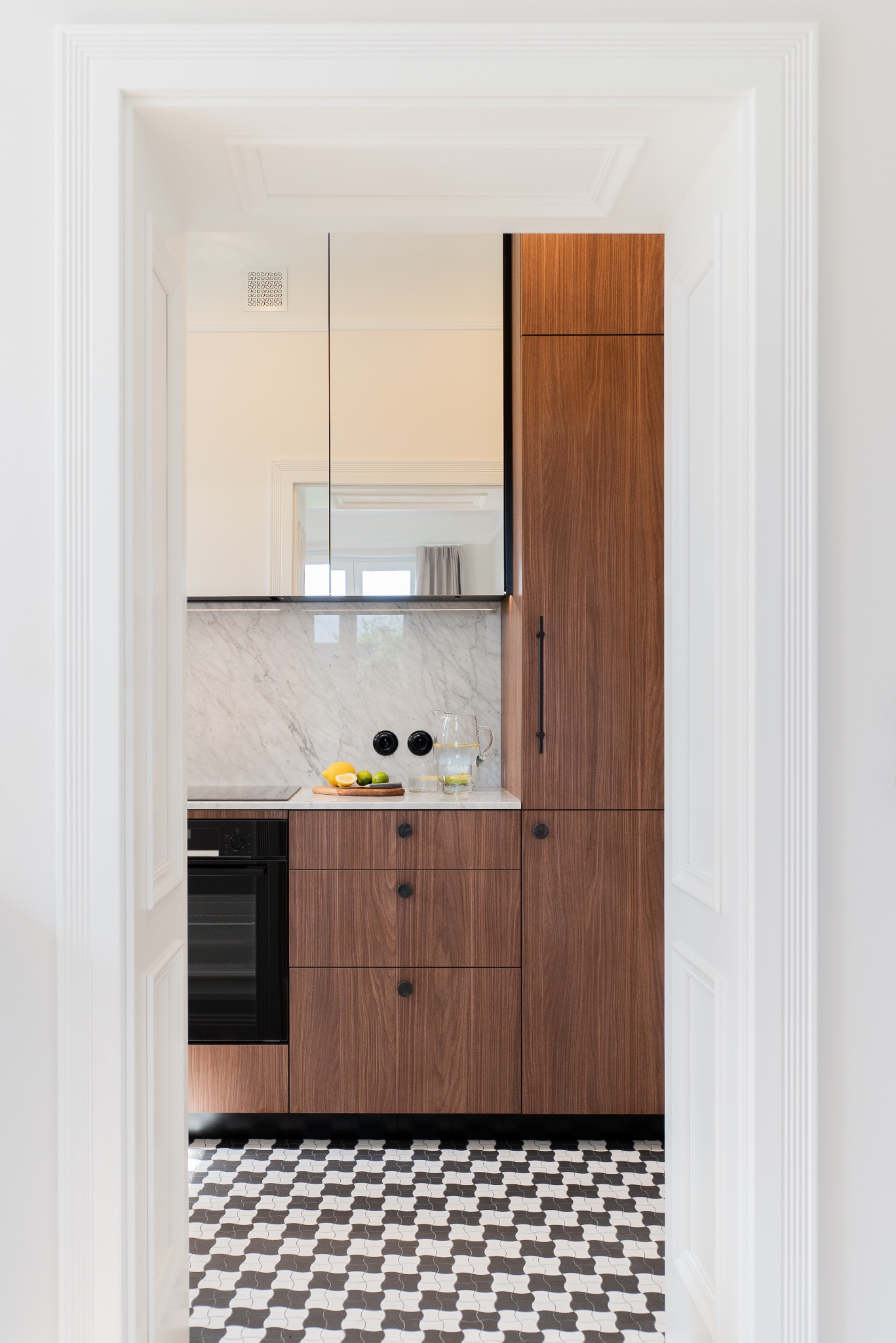 Nowoczesna kuchnia z drewnianymi szafkami, marmurowym blatem i czarno-białą podłogą w kratkę, widziana przez otwarte, białe drzwi z dekoracyjnymi listwami.