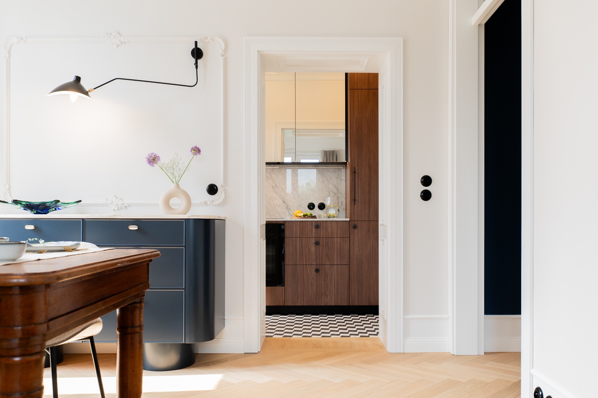 Eleganckie wnętrze: widok na kuchnię z drewnianymi szafkami, marmurowym blatem i czarno-białą podłogą w jodełkę, z salonu z ciemnoniebieską komodą, drewnianym stołem i kwiatami w wazonie.