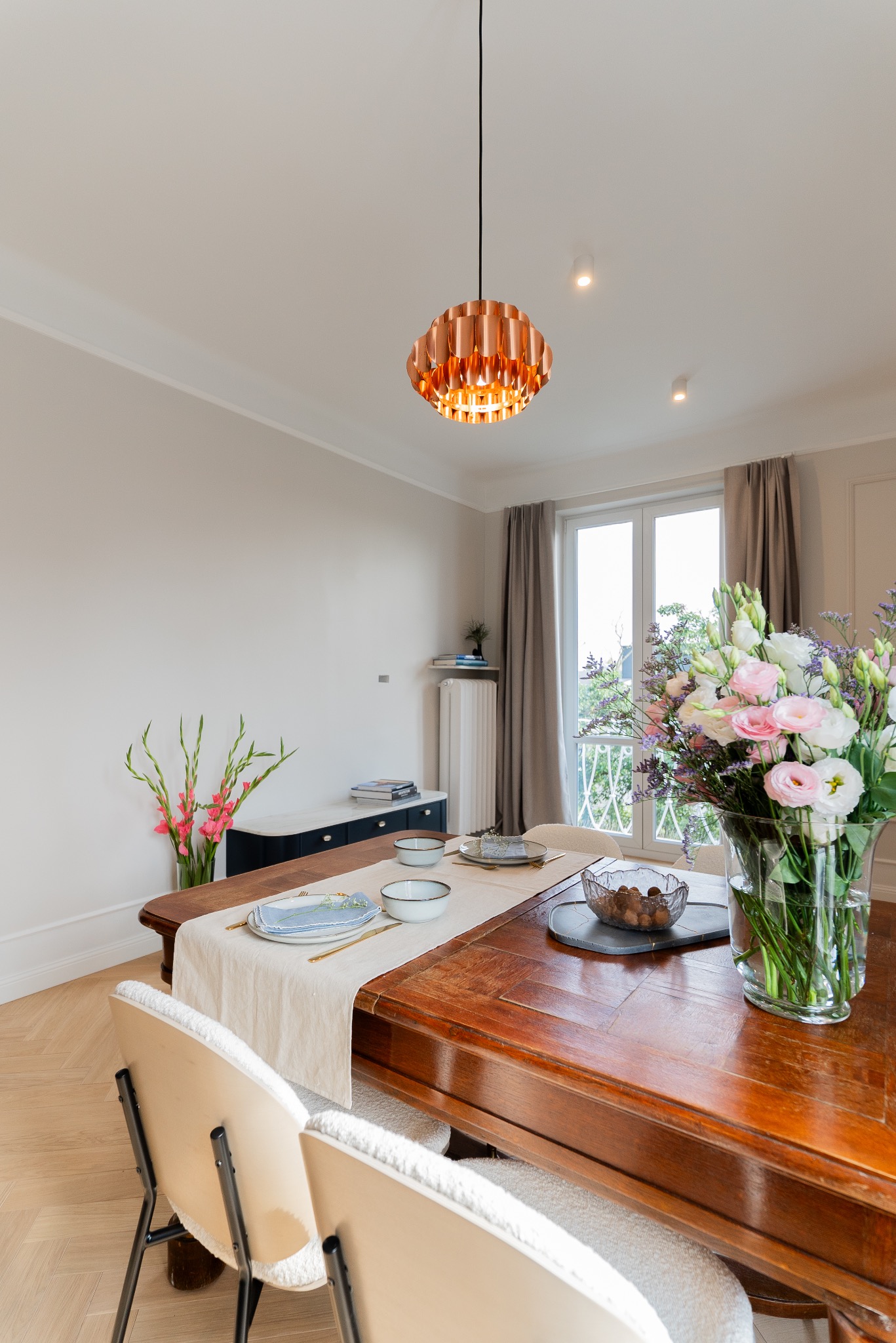 Jadalnia w jasnych barwach z drewnianym stołem nakrytym do posiłku, miedziana lampa wisząca, bukiet kwiatów w wazonie i widok z okna na zieleń.