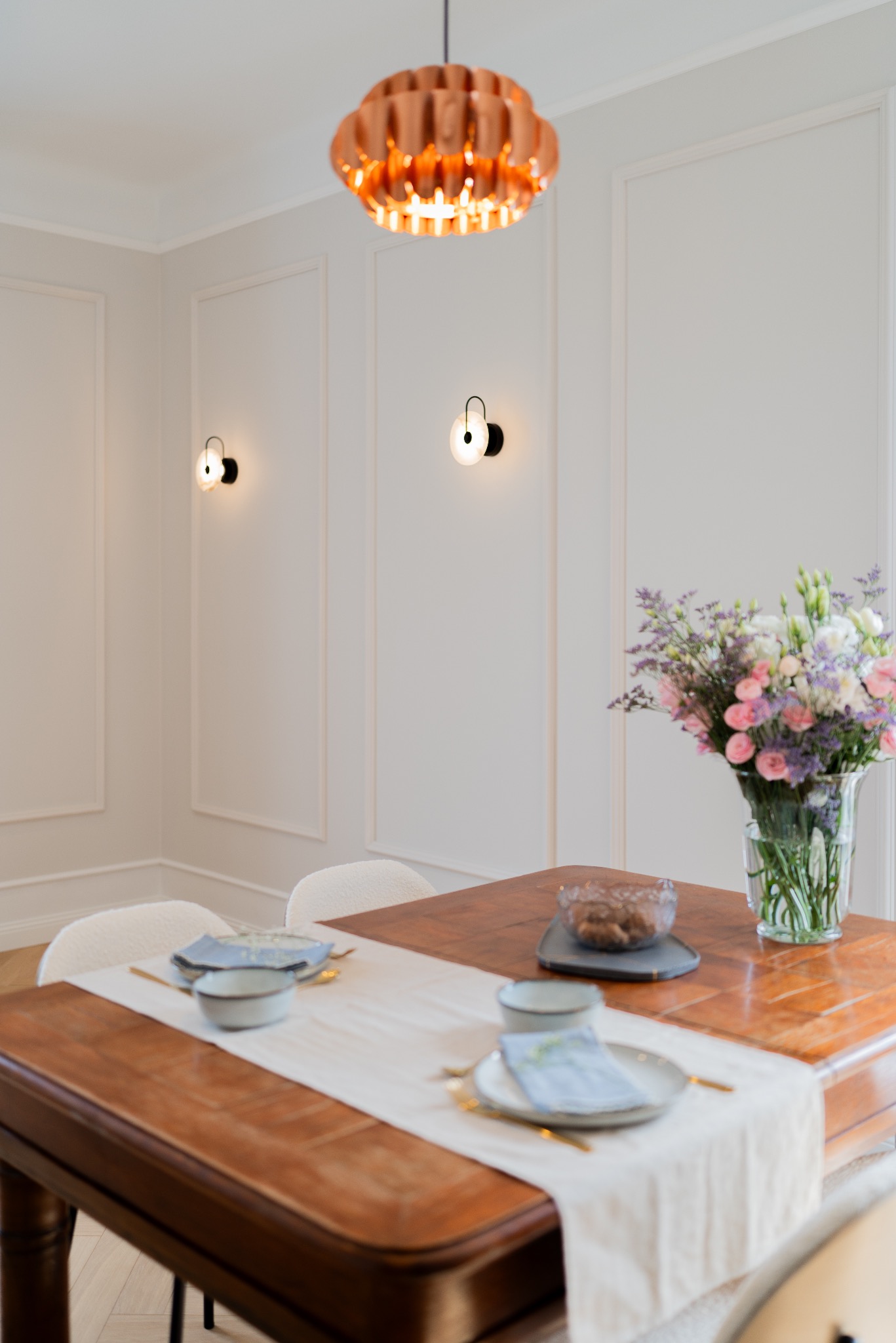 Elegancka jadalnia z drewnianym stołem, nakrytym dla dwóch osób, z bukietem kwiatów w wazonie. Na ścianach dekoracyjne panele i nowoczesne kinkiety, a nad stołem miedziana lampa.