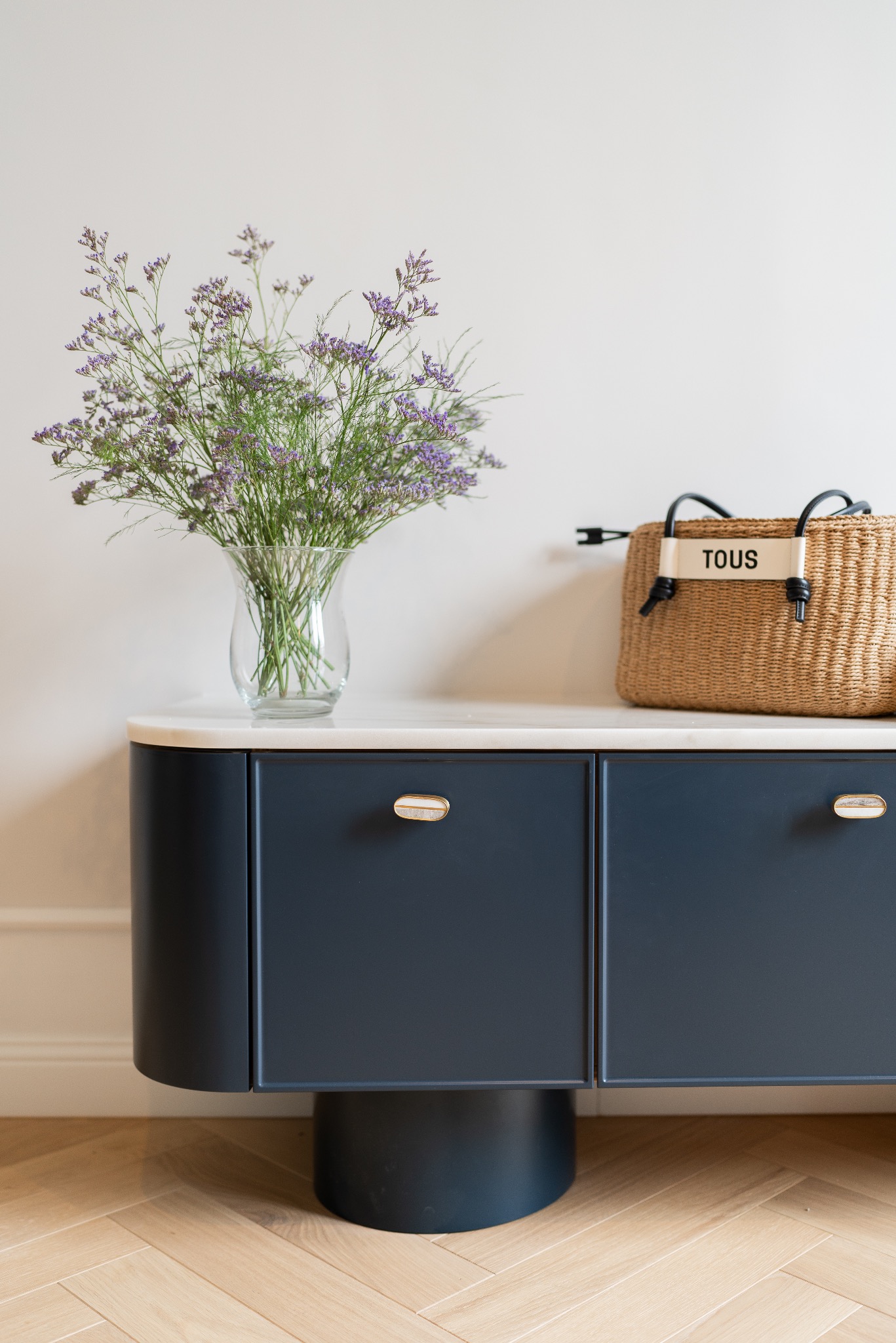 Stylowa, ciemnoniebieska komoda z marmurowym blatem, na którym stoi szklany wazon z fioletowymi kwiatami i słomkowa torebka marki TOUS.