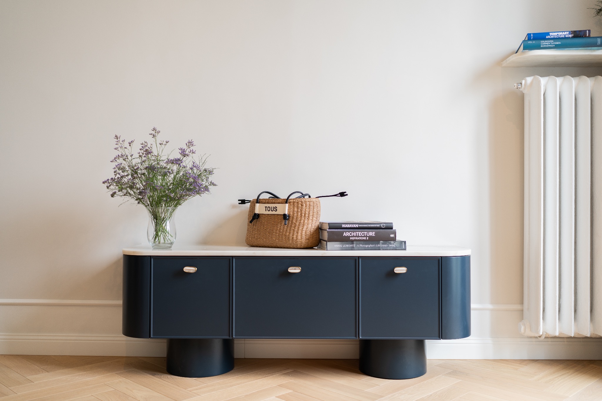 Granatowa komoda z białym blatem, okrągłymi nóżkami i złotymi uchwytami, na blacie wazon z fioletowymi kwiatami, słomkowa torba Tous i stos książek o architekturze, na ścianie biały grzejnik.