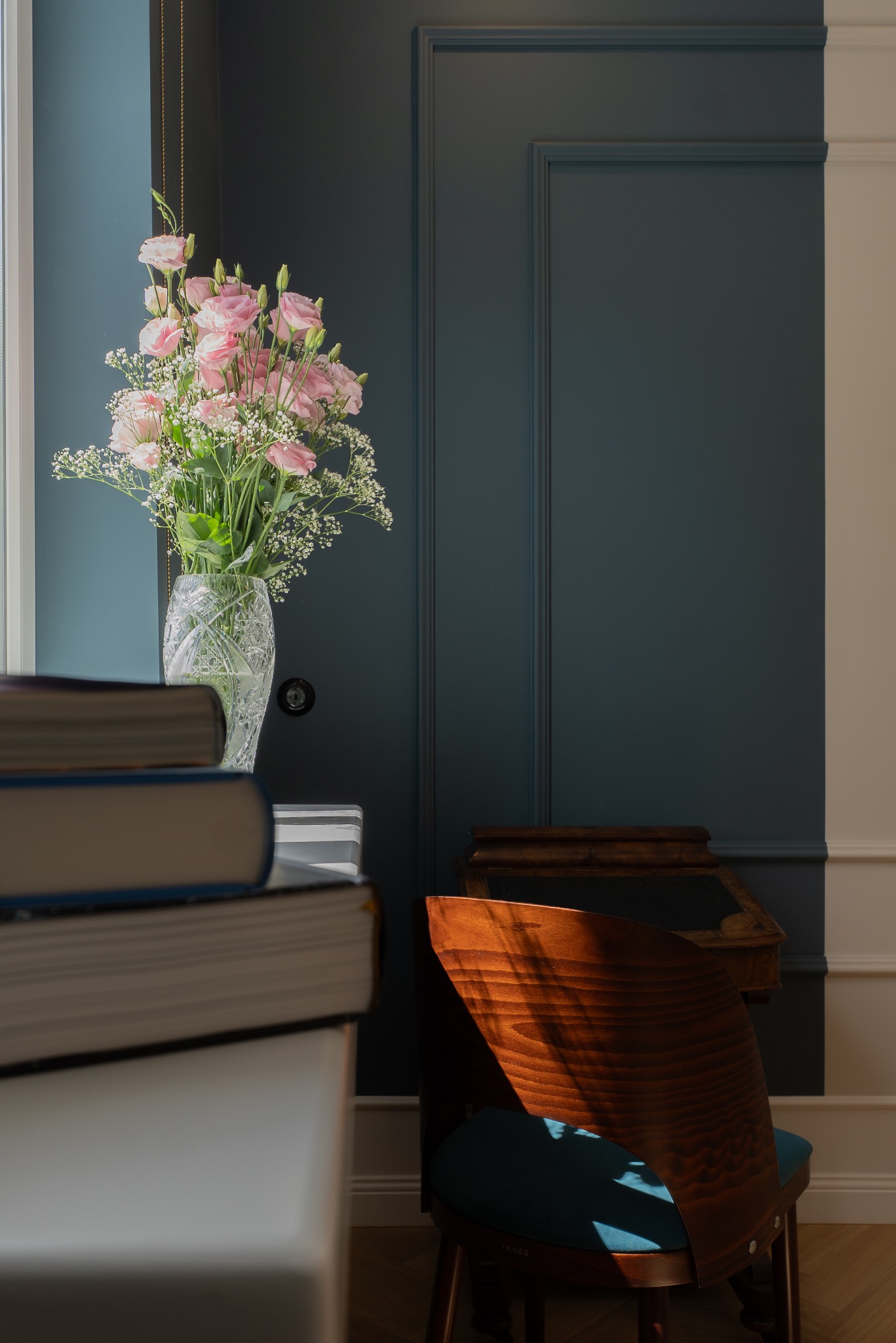 Kryształowy wazon z bukietem różowych kwiatów i gipsówki na tle granatowej ściany z dekoracyjnymi listwami, obok stos książek i drewniane krzesło w promieniach słońca.