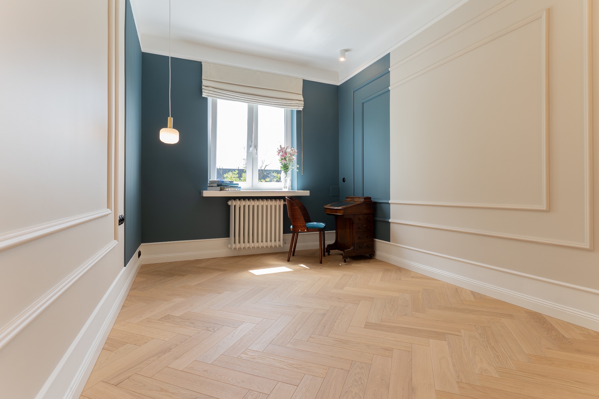 Jasny parkiet w jodełkę, niebieska ściana z białymi sztukateriami, drewniane biurko i krzesło z niebieskim siedziskiem przy oknie z grzejnikiem i książkami.
