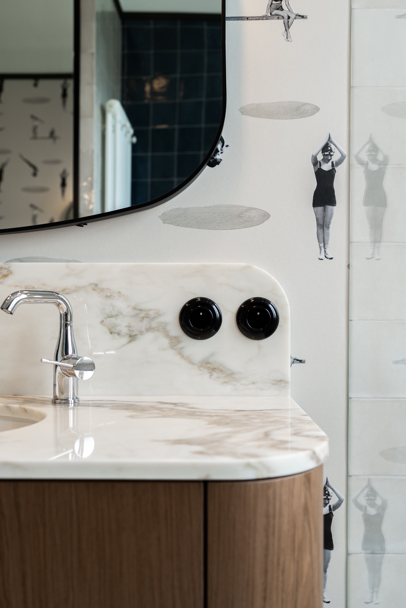 Umywalka z marmurowym blatem, chromowaną baterią i dwoma czarnymi włącznikami, na tle ściany z motywem pływaczek w stylu retro.