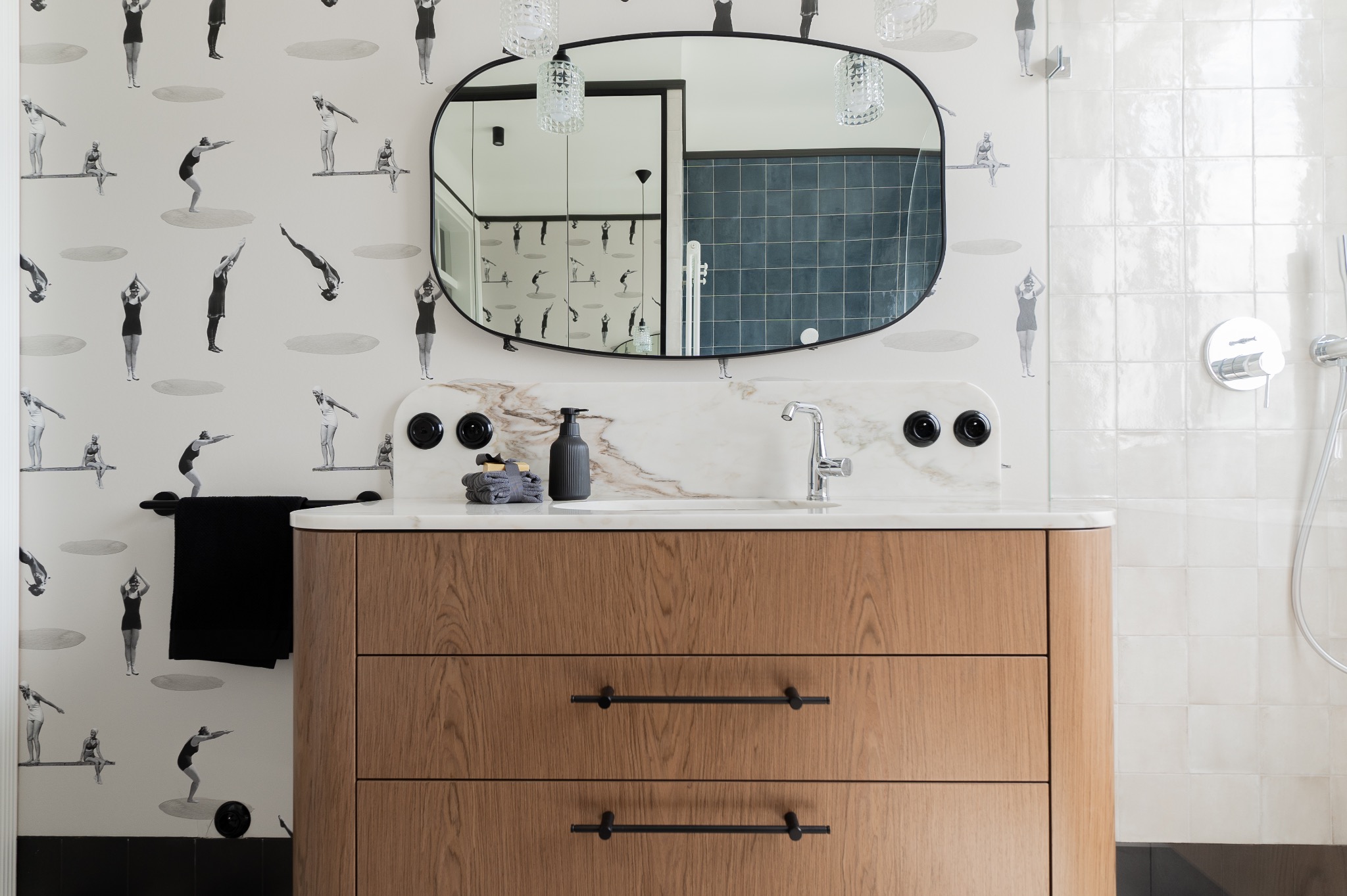 Nowoczesna łazienka z drewnianą szafką pod umywalką, marmurowym blatem, owalnym lustrem i tapetą w stylu retro z motywem pływaczek.