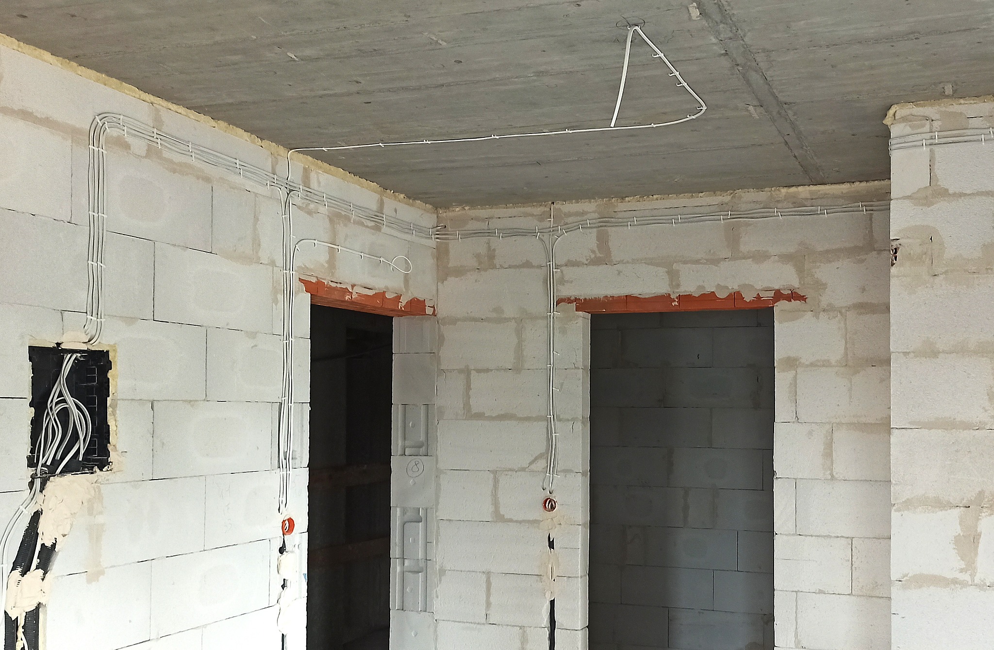 Surowy stan: białe ściany z betonu komórkowego, widoczne kable elektryczne prowadzone po ścianach i suficie, puszki instalacyjne. Instalacja w nowym budynku.