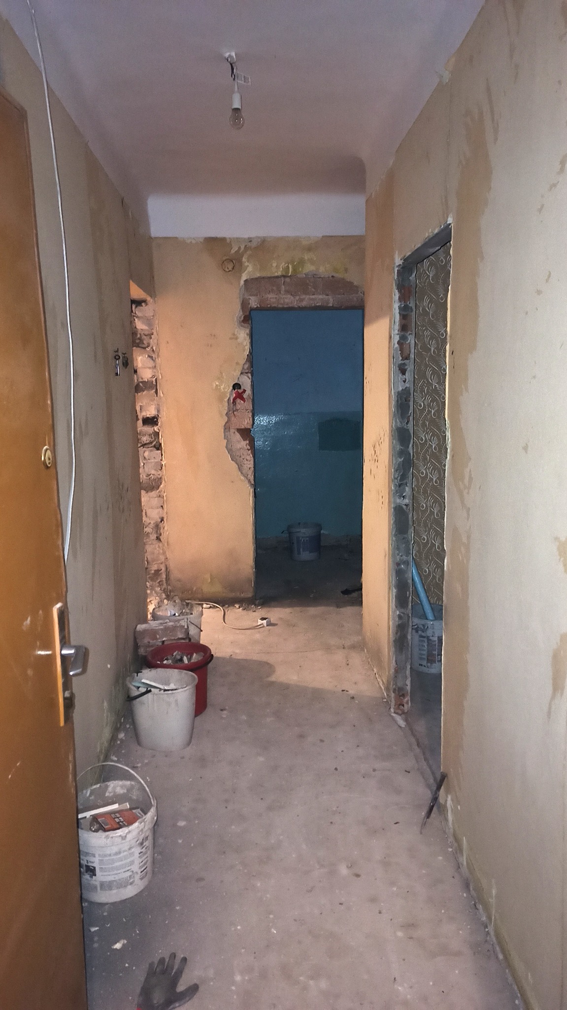 Korytarz w trakcie remontu w Dąbrowie Górniczej. Widoczne ściany przygotowane do malowania, fragmenty odsłoniętej cegły, wiadra z farbą i goła żarówka.