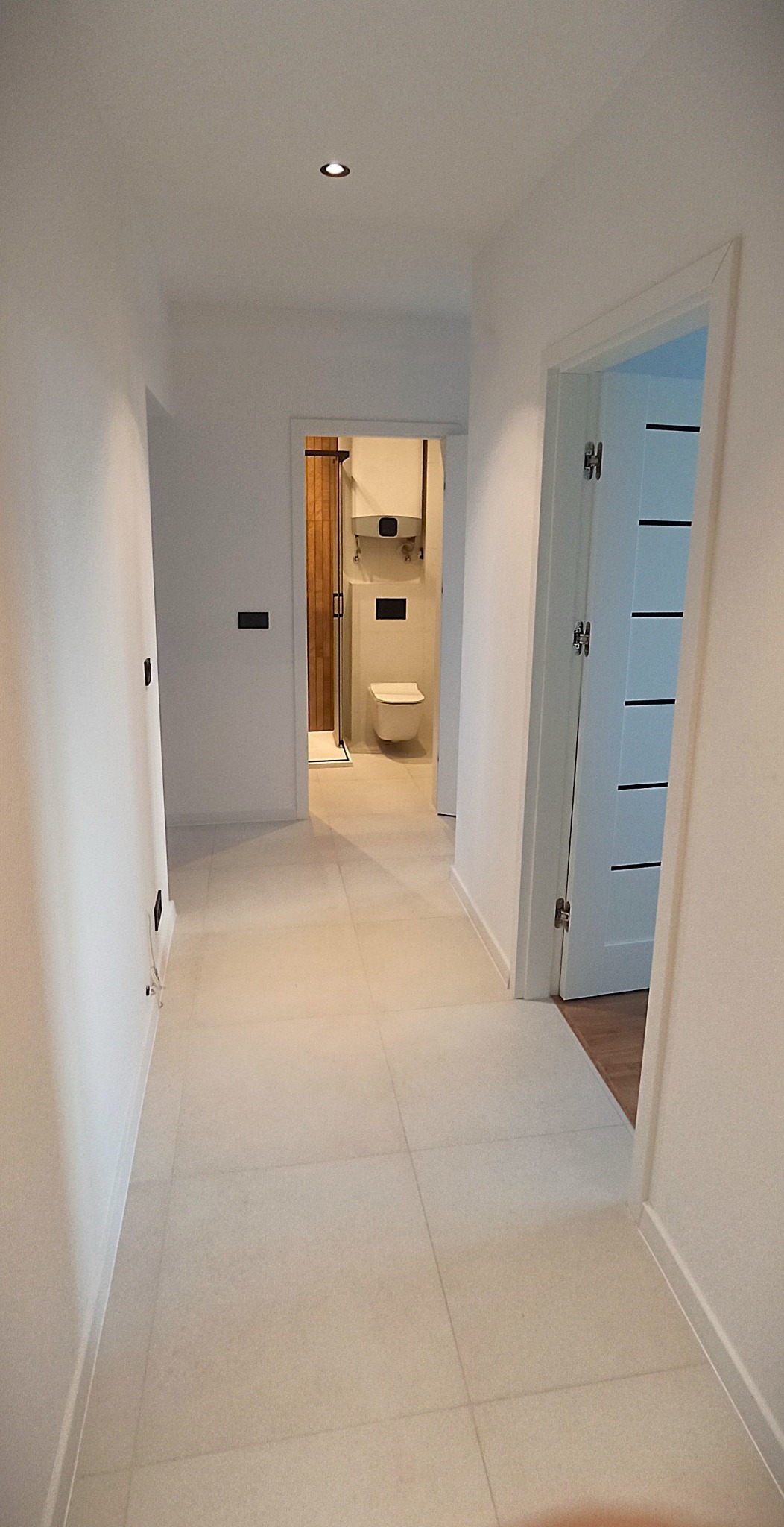 Jasny korytarz z beżowymi płytkami podłogowymi, prowadzący do łazienki z kabiną prysznicową i toaletą. Widoczne białe ściany i drzwi z czarnymi akcentami.