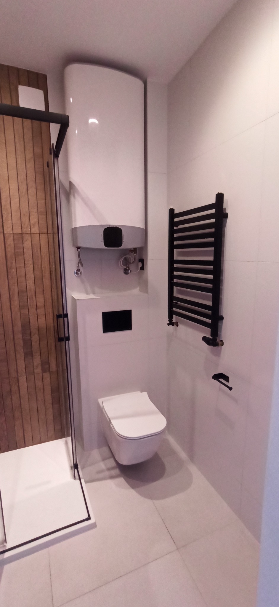 Nowoczesna łazienka z kabiną prysznicową z drewnianymi panelami, podwieszaną toaletą i grzejnikiem drabinkowym w czarnym kolorze. Minimalistyczny design, jasne płytki.