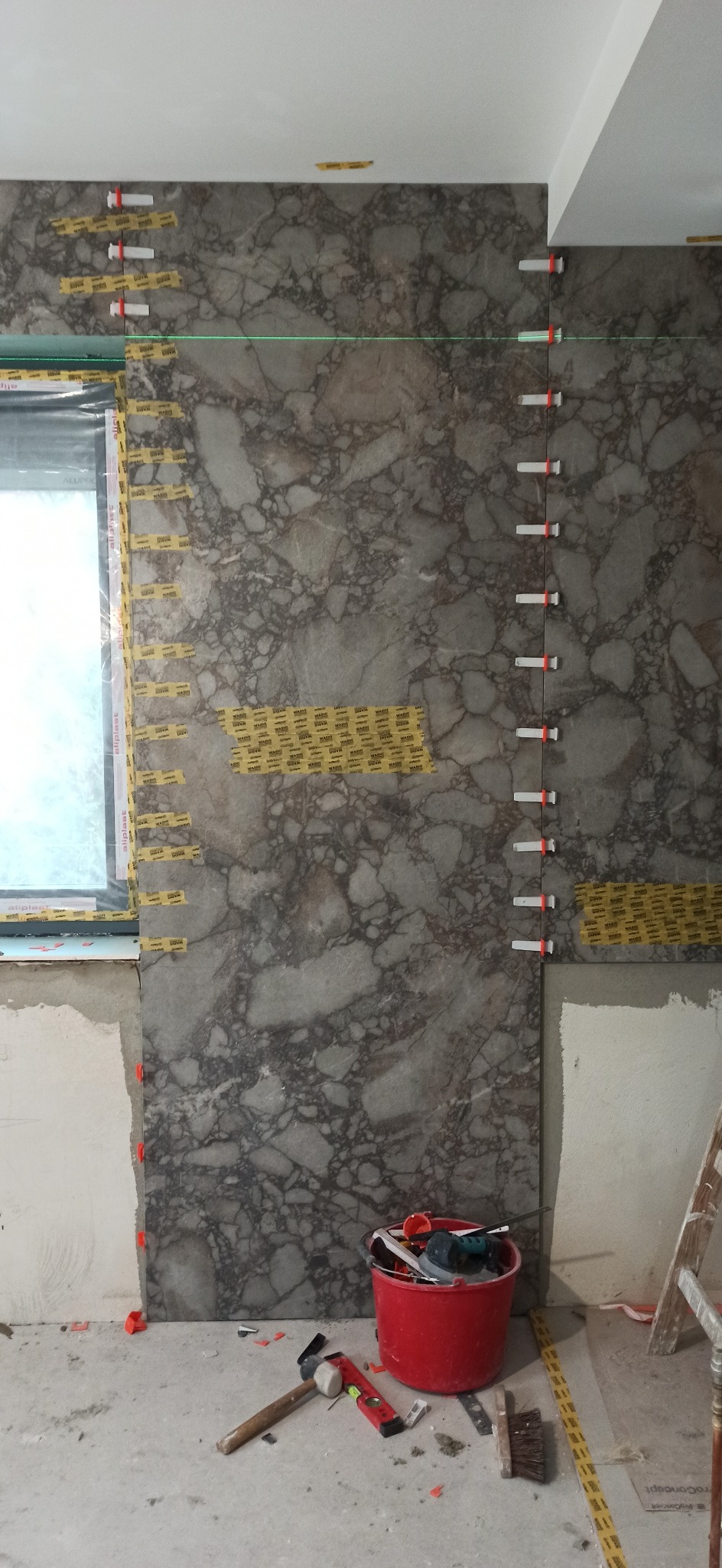 Montaż płytek imitujących marmur na ścianie, widoczne narzędzia i poziomica laserowa. Etap prac wykończeniowych w pomieszczeniu z oknem.