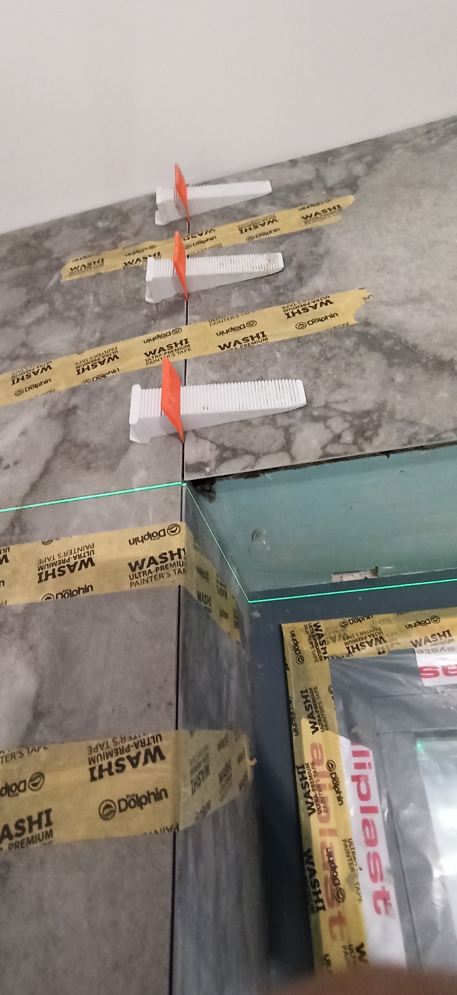 Precyzyjne układanie płytek z użyciem poziomicy laserowej, klipsów i taśmy malarskiej. Detal wykończenia w trakcie prac. Szare płytki imitujące marmur.