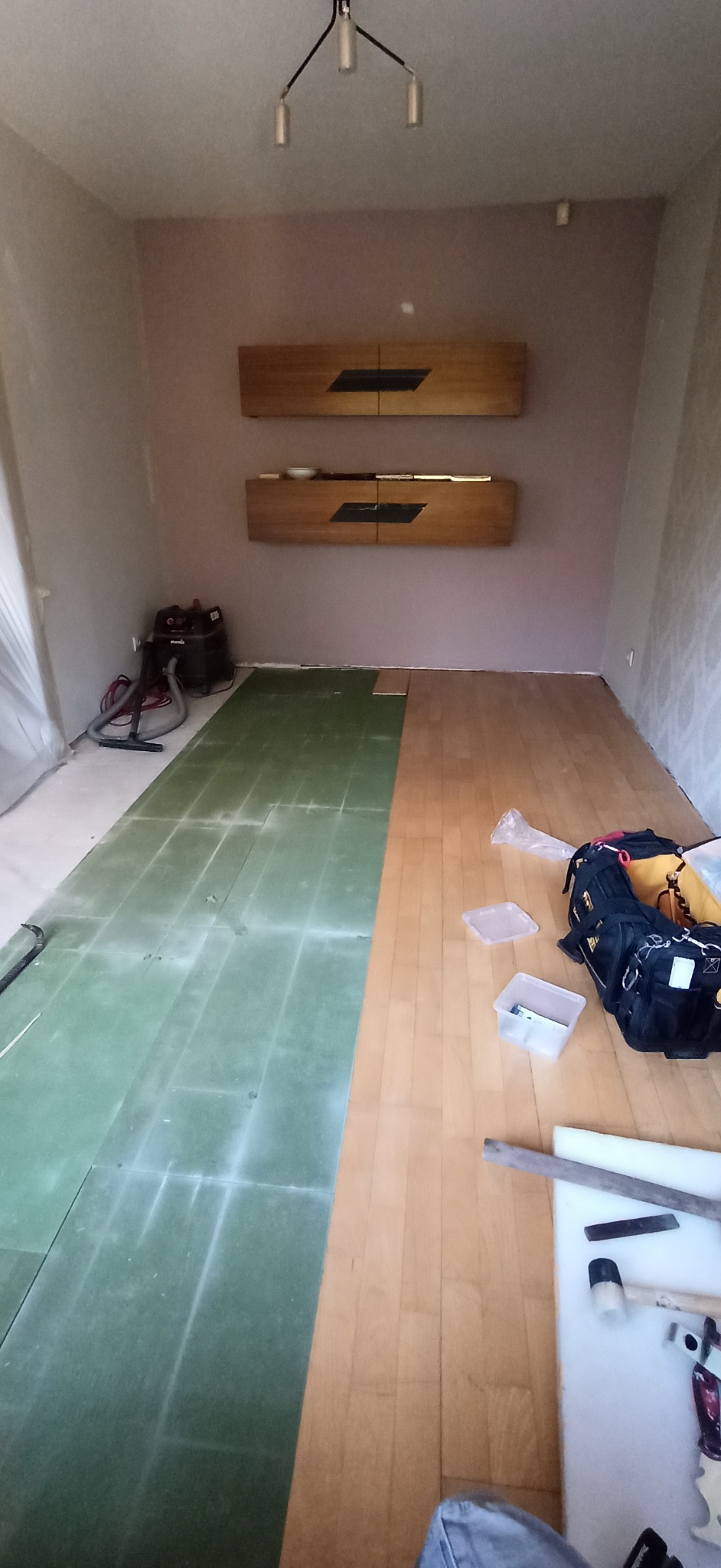 Pokój w trakcie układania paneli podłogowych. Część podłogi pokryta zielonym podkładem, część panelami. Widoczne narzędzia i torba monterska.