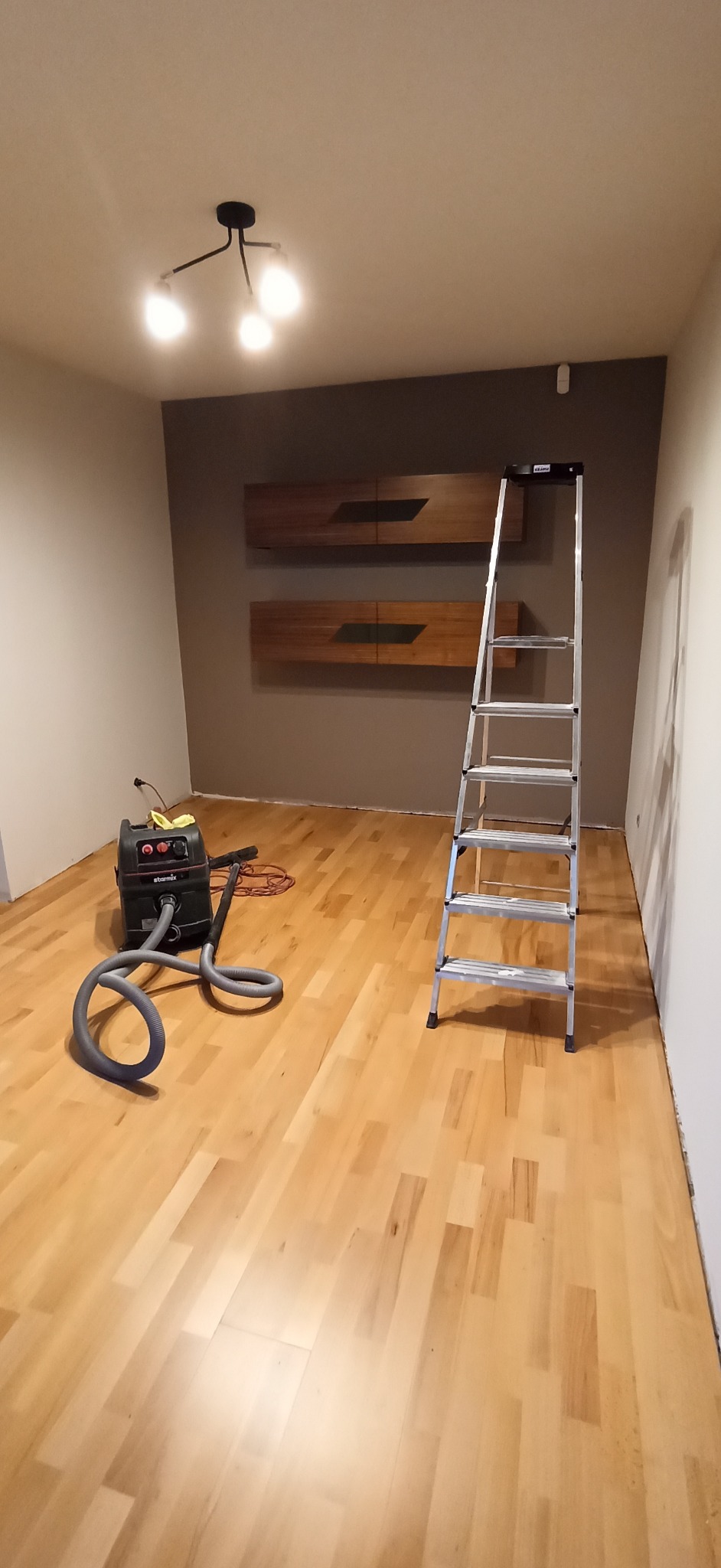 Pokój po remoncie: jasna podłoga z paneli, drabina oparta o brązową ścianę z szafkami, odkurzacz przemysłowy. Widok na wnętrze po pracach wykończeniowych.