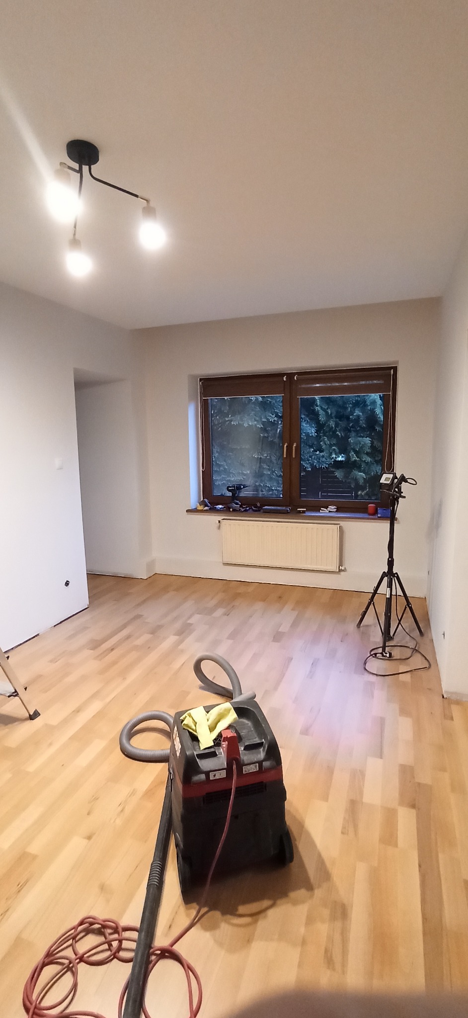 Jasne panele podłogowe po cyklinowaniu, widoczny odkurzacz przemysłowy i lampa na statywie. Świeżo wyremontowane wnętrze z oknem i grzejnikiem.