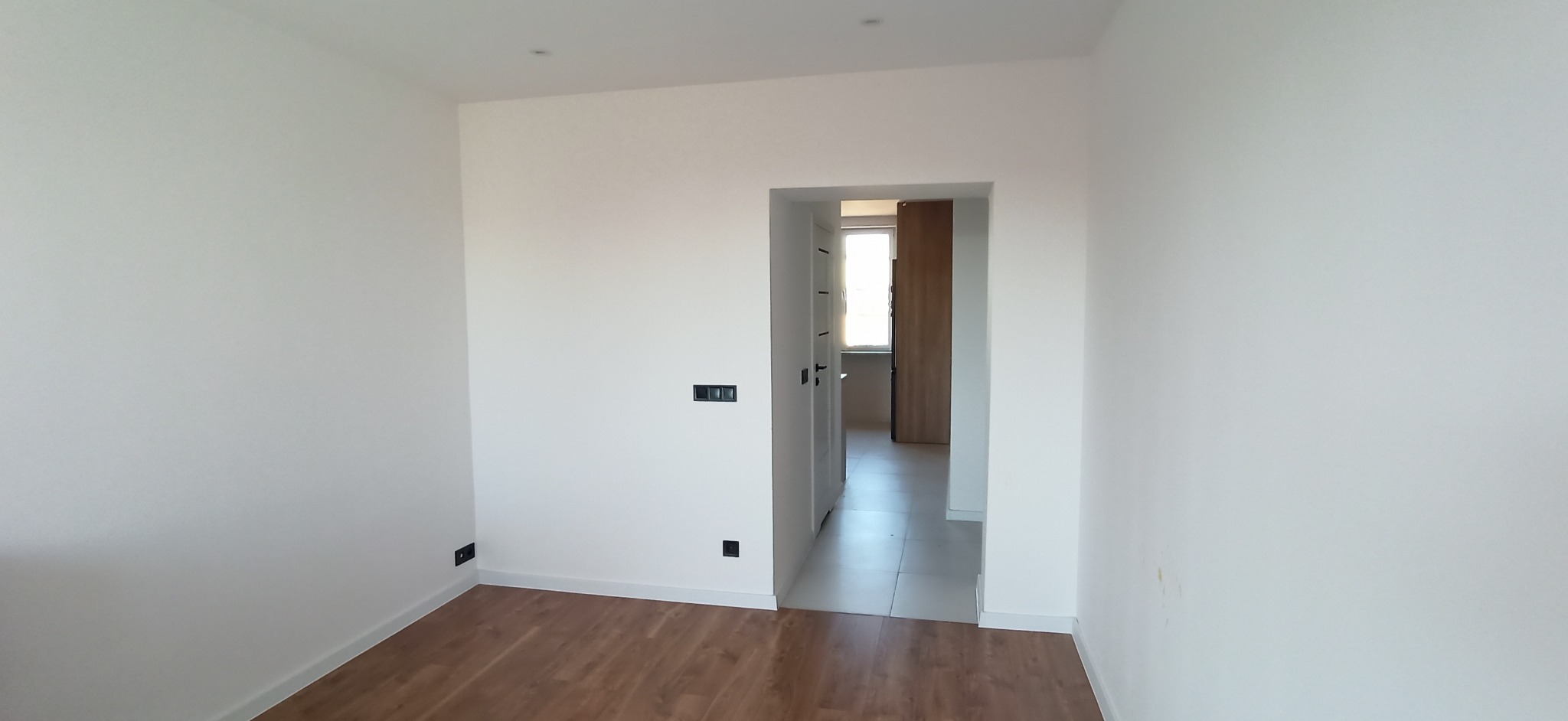 Jasne wnętrze po remoncie: świeżo pomalowane białe ściany, ułożone panele podłogowe w odcieniach brązu, widoczne gniazdka elektryczne i drzwi do kolejnego pomieszczenia.