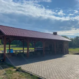 Zadaszona wiata z ceglanym grillem, stołami i ławkami na balach słomy, przygotowana do imprezy plenerowej, widok na zieloną okolicę i błękitne niebo z chmurami.