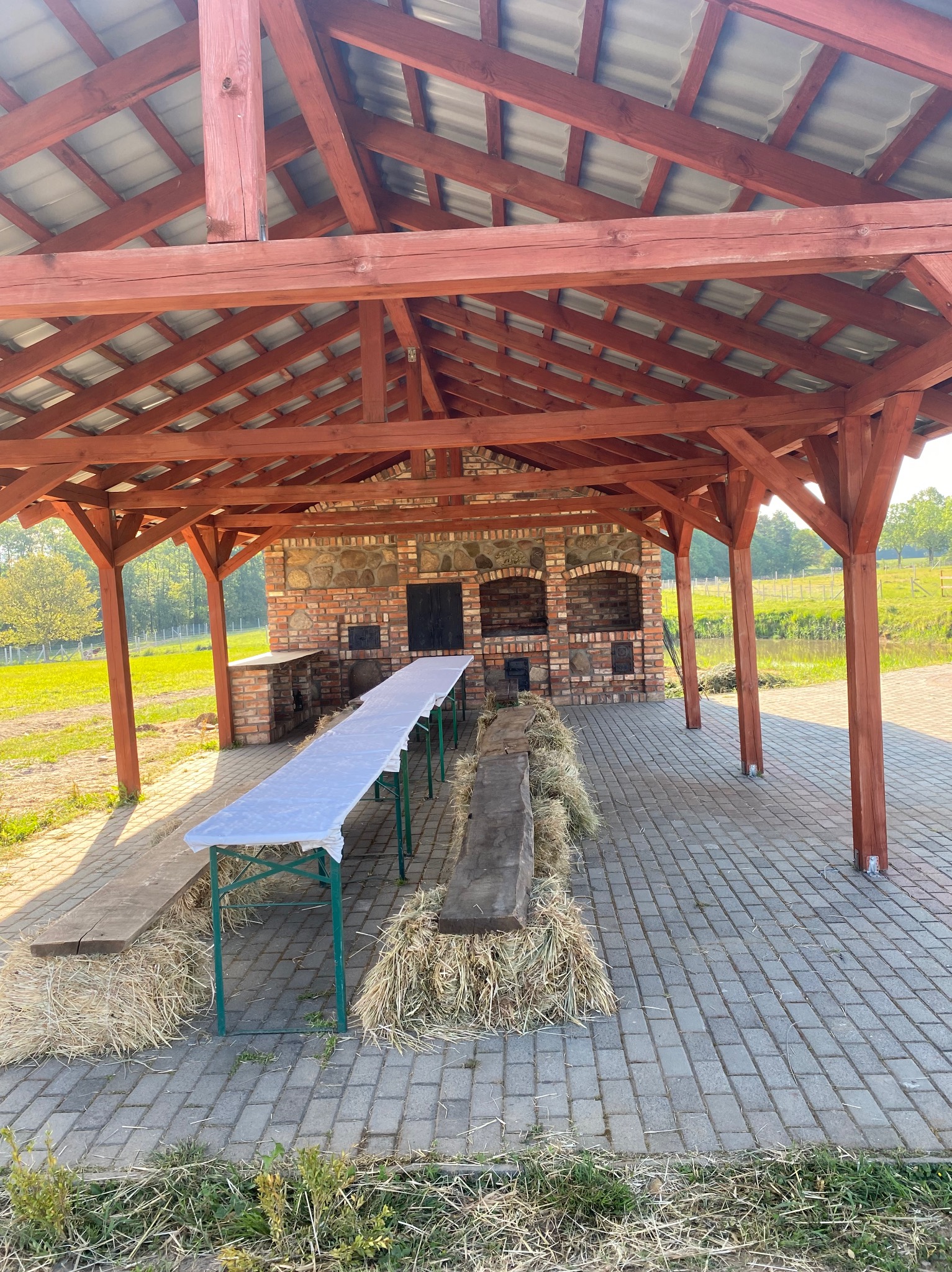 Zadaszona wiata z ceglanym grillem, długim stołem przykrytym białym obrusem i ławkami na belach słomy, przygotowana na imprezę plenerową.