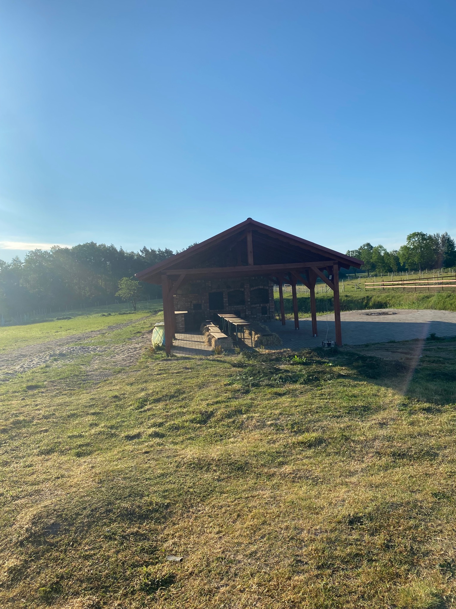 Zadaszona altana z ceglanym grillem i drewnianymi ławami na terenie zielonym, oświetlona porannym słońcem.