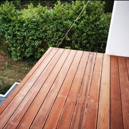 Rad-Bud usługi remontowo budowlane - Nowoczesny taras z desek kompozytowych w odcieniach brązu, widok z góry na tle zielonego żywopłotu i białej ściany budynku.