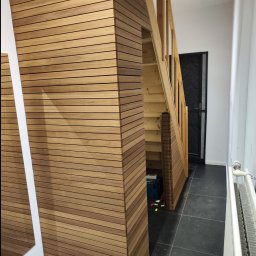 Rad-Bud usługi remontowo budowlane - Korytarz z drewnianą boazerią na ścianie i schodami, widoczna skrzynka z narzędziami pod schodami, czarne płytki na podłodze.