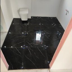 Rad-Bud usługi remontowo budowlane - Łazienka w trakcie prac: ułożone czarne płytki podłogowe z białym żyłkowaniem, biała wisząca toaleta, białe ściany, widoczne krzyżyki dystansowe i poziomica laserowa.