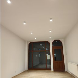 Rad-Bud usługi remontowo budowlane - Puste, świeżo wykończone wnętrze z panelami podłogowymi imitującymi drewno, białymi ścianami, oknem i drzwiami z brązowymi ramami oraz zamontowanym grzejnikiem.