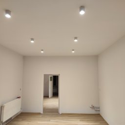 Rad-Bud usługi remontowo budowlane - Pomalowane na biało puste pomieszczenie z panelami podłogowymi i białym grzejnikiem, oświetlone punktowymi lampami sufitowymi, z widokiem na otwarte drzwi do kolejnego pomieszczenia.