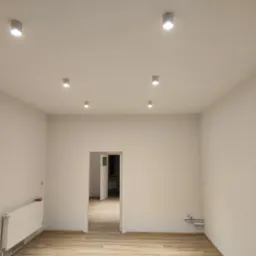 Pomalowane na biało puste pomieszczenie z panelami podłogowymi i białym grzejnikiem, oświetlone punktowymi lampami sufitowymi, z widokiem na otwarte drzwi do kolejnego pomieszczenia.