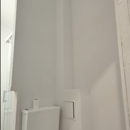 Rad-Bud usługi remontowo budowlane - Biała, świeżo pomalowana ściana w łazience z zamontowaną toaletą i widoczną kabiną prysznicową w tle.