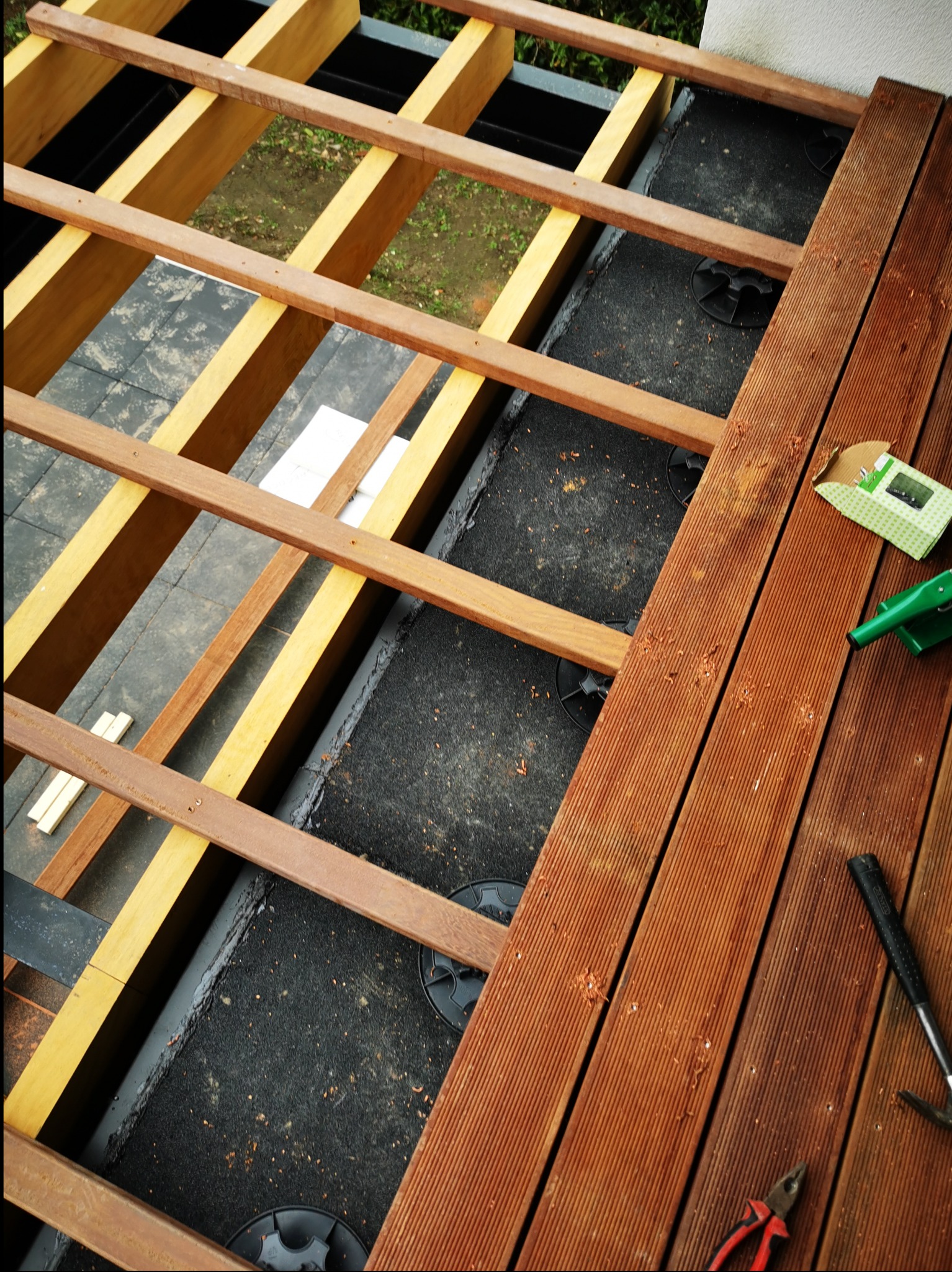 Montaż drewnianego tarasu na regulowanych wspornikach, widoczne legary, deski tarasowe, narzędzia i opakowanie po wkrętach.