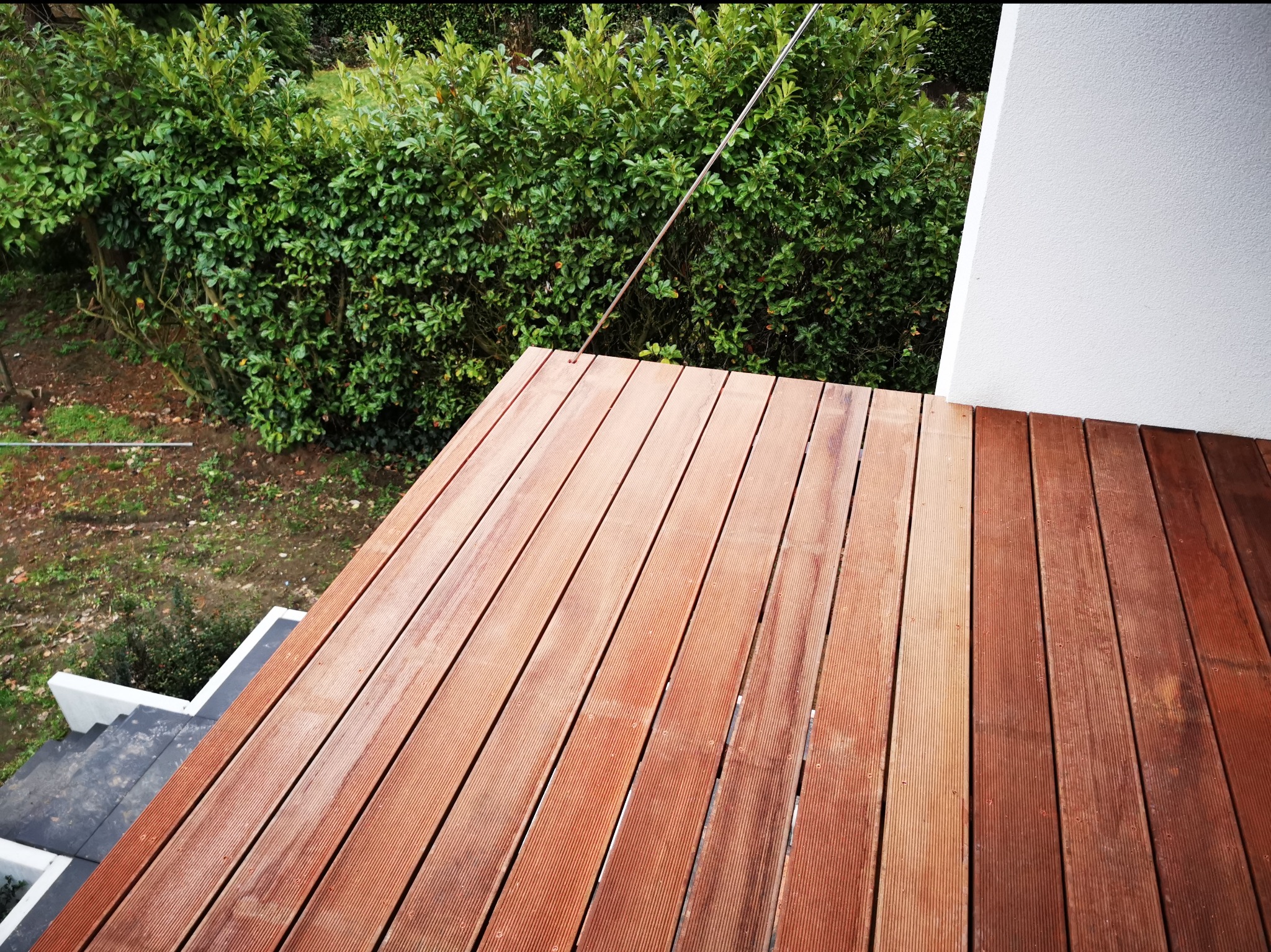 Nowoczesny taras z desek kompozytowych w odcieniach brązu, widok z góry na tle zielonego żywopłotu i białej ściany budynku.