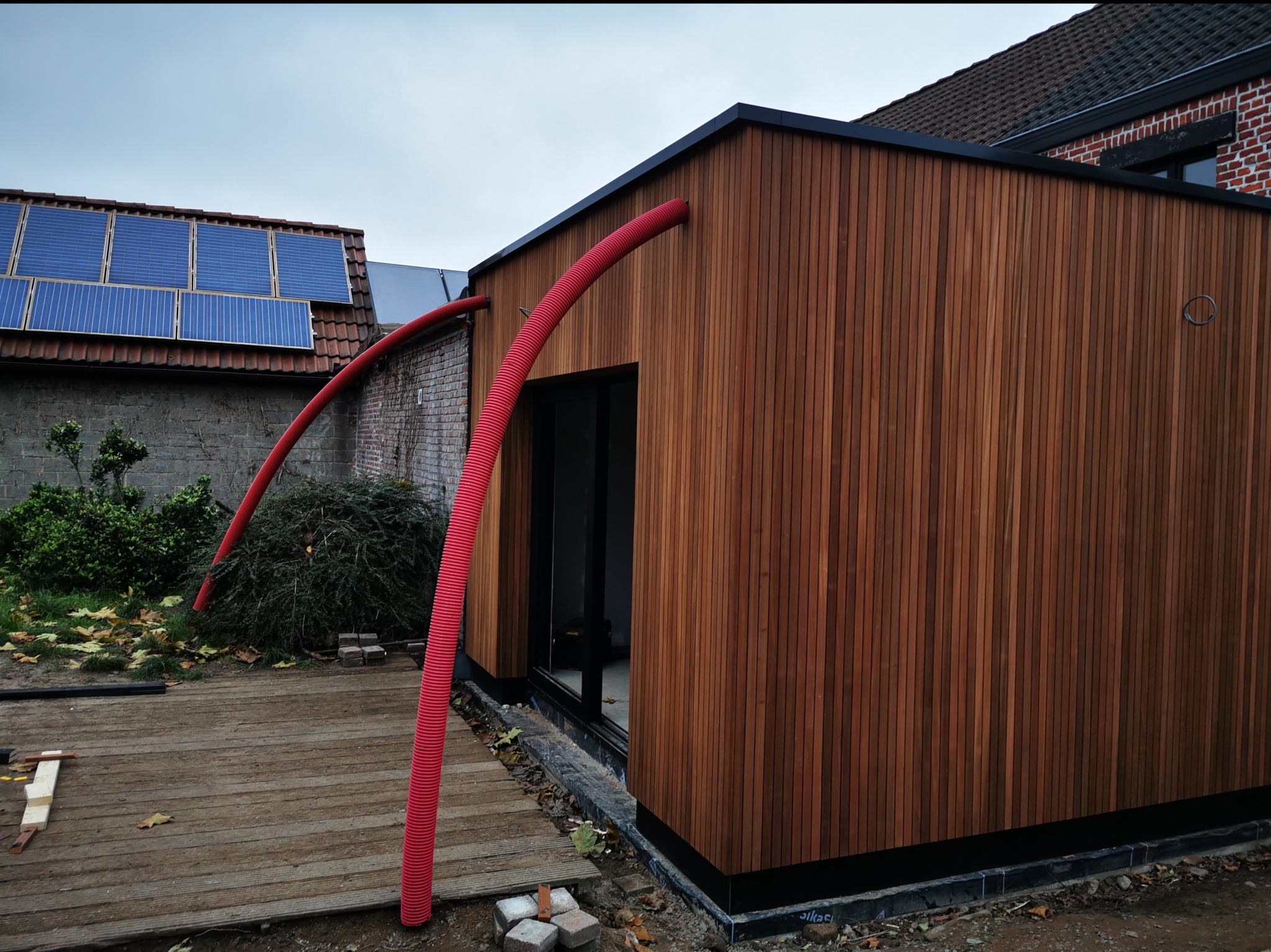 Nowoczesna elewacja budynku wykończona drewnianymi panelami, z widocznymi czerwonymi rurami wentylacyjnymi na zewnątrz i drewnianym tarasem w trakcie budowy.