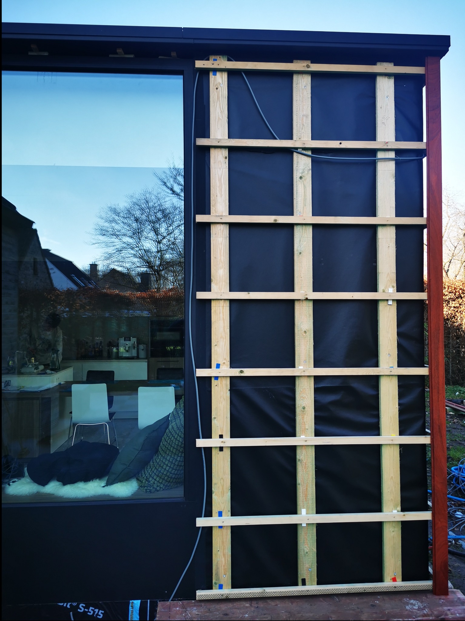 Montaż drewnianej konstrukcji na czarnej membranie elewacyjnej, widoczny kabel i fragment wnętrza przez okno.