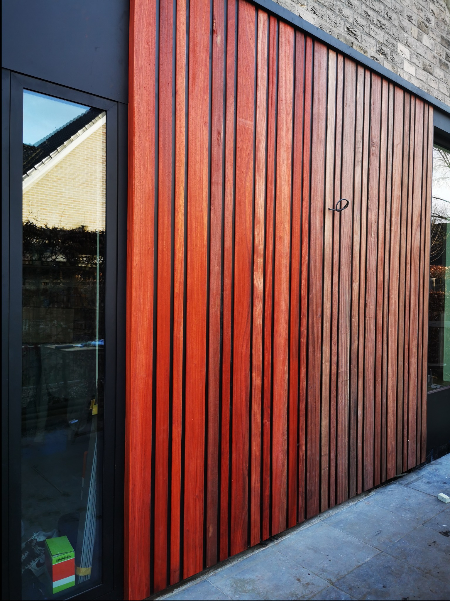 Nowoczesna elewacja z pionowych, drewnianych paneli o ciepłym, czerwono-brązowym odcieniu, z czarnymi wstawkami i ramą okienną, widoczna z zewnątrz na tle szarej kostki brukowej.