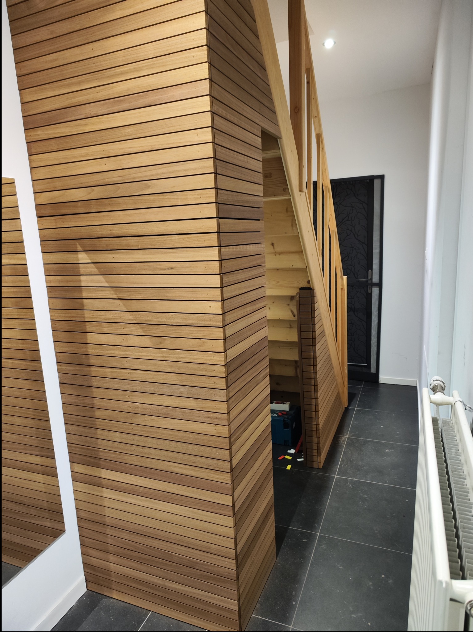 Korytarz z drewnianą boazerią na ścianie i schodami, widoczna skrzynka z narzędziami pod schodami, czarne płytki na podłodze.