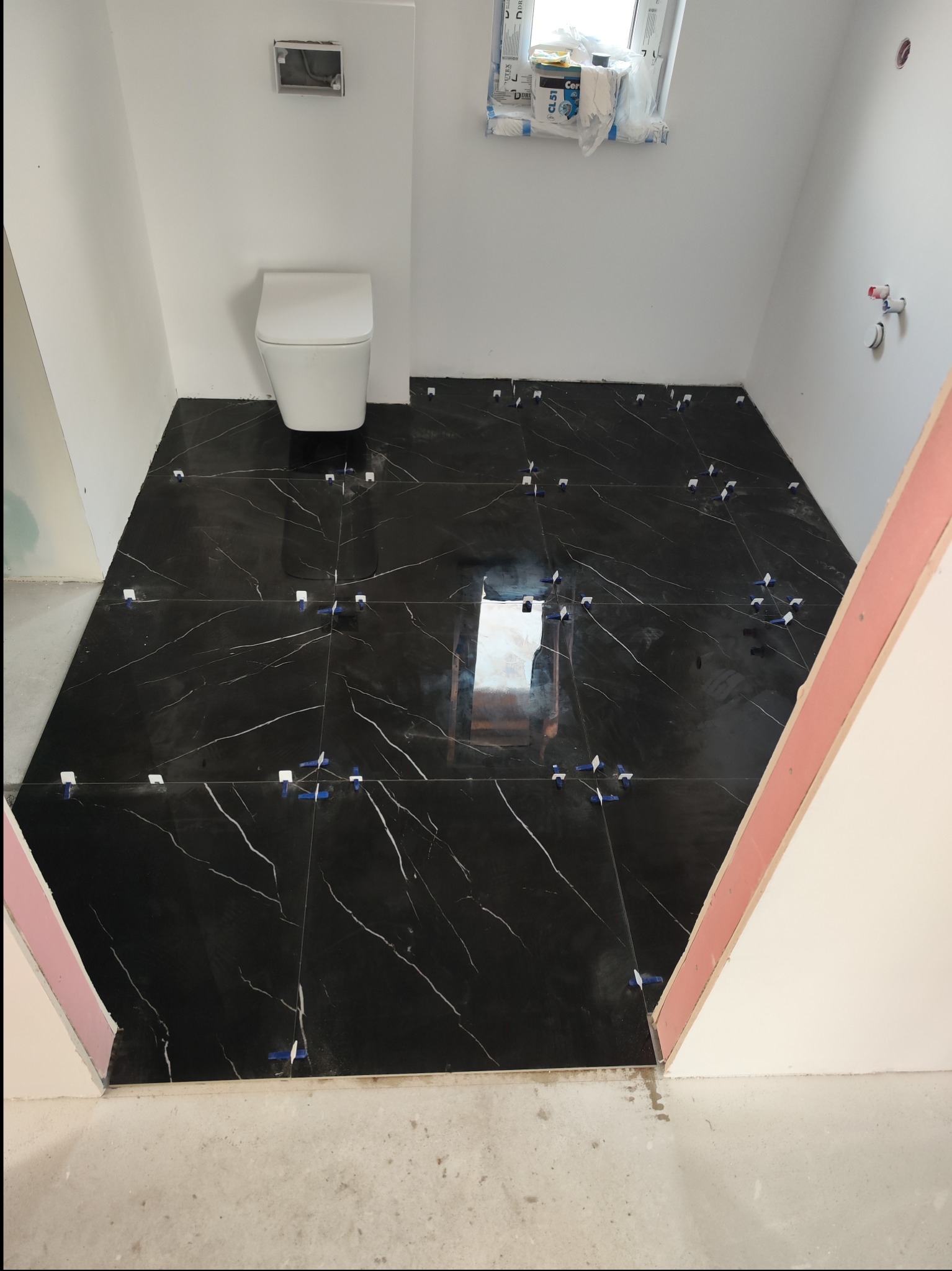 Łazienka w trakcie prac: ułożone czarne płytki podłogowe z białym żyłkowaniem, biała wisząca toaleta, białe ściany, widoczne krzyżyki dystansowe i poziomica laserowa.