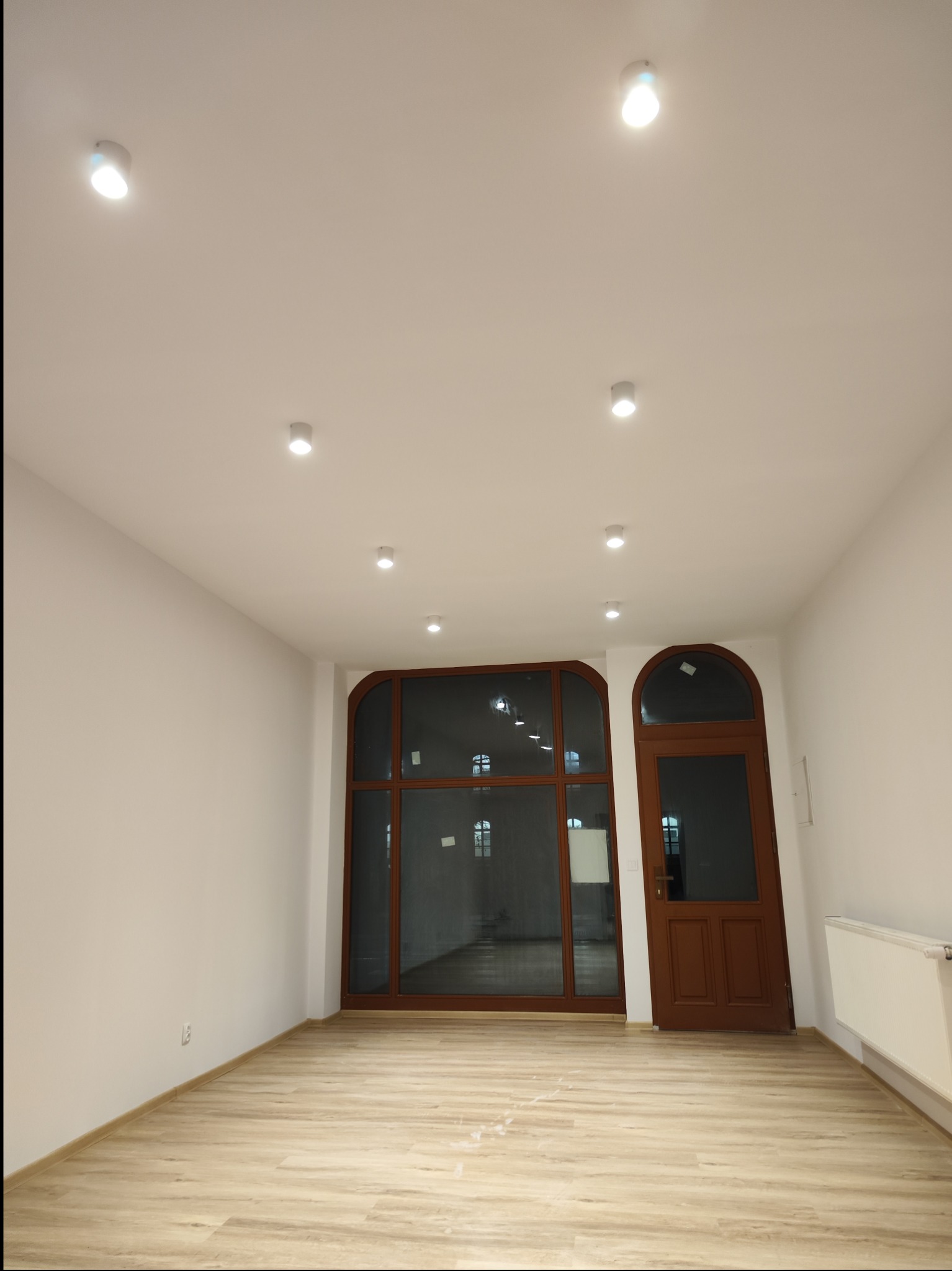 Puste, świeżo wykończone wnętrze z panelami podłogowymi imitującymi drewno, białymi ścianami, oknem i drzwiami z brązowymi ramami oraz zamontowanym grzejnikiem.