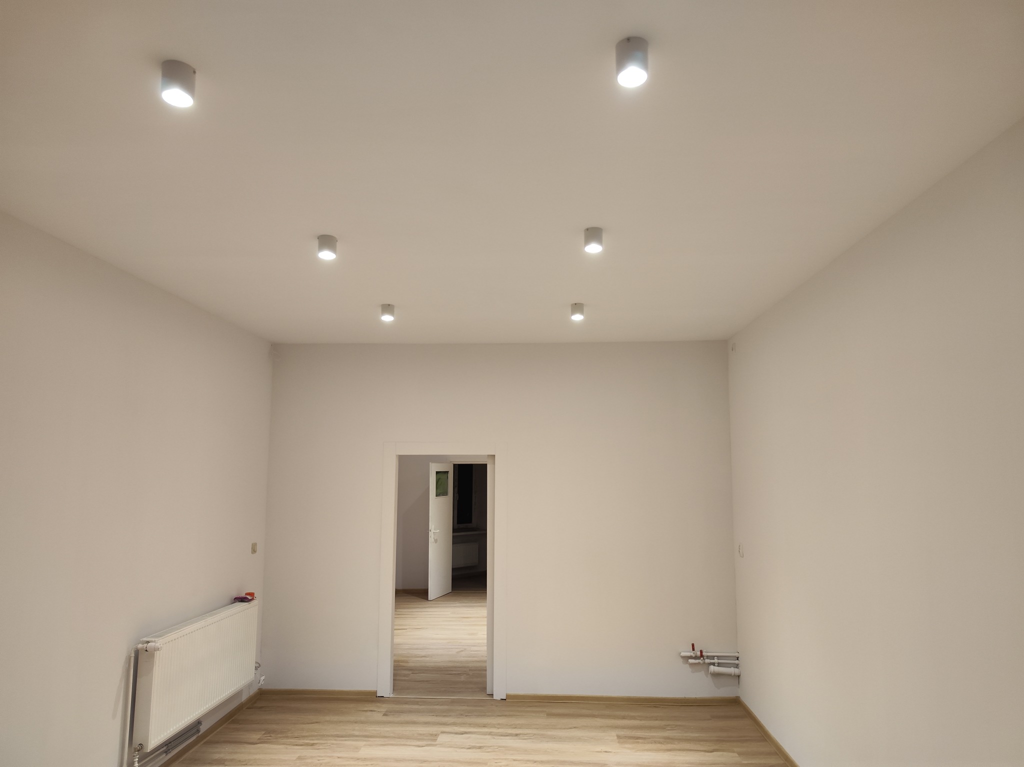 Pomalowane na biało puste pomieszczenie z panelami podłogowymi i białym grzejnikiem, oświetlone punktowymi lampami sufitowymi, z widokiem na otwarte drzwi do kolejnego pomieszczenia.
