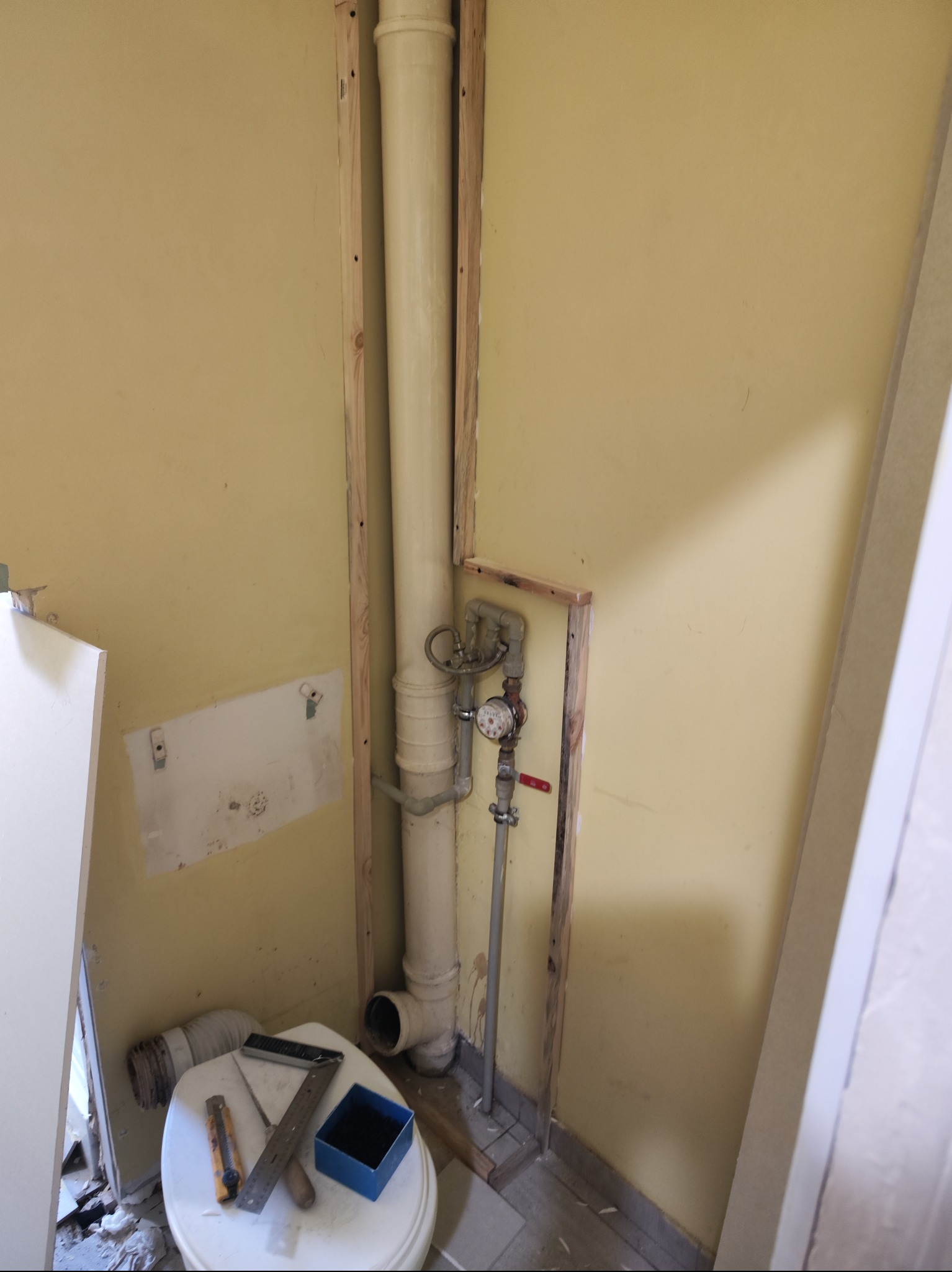 Żółta ściana z odsłoniętą rurą kanalizacyjną w drewnianej ramie, na sedesie nóż tapicerski, kątownik i niebieskie pudełko, fragment demontażu obudowy.