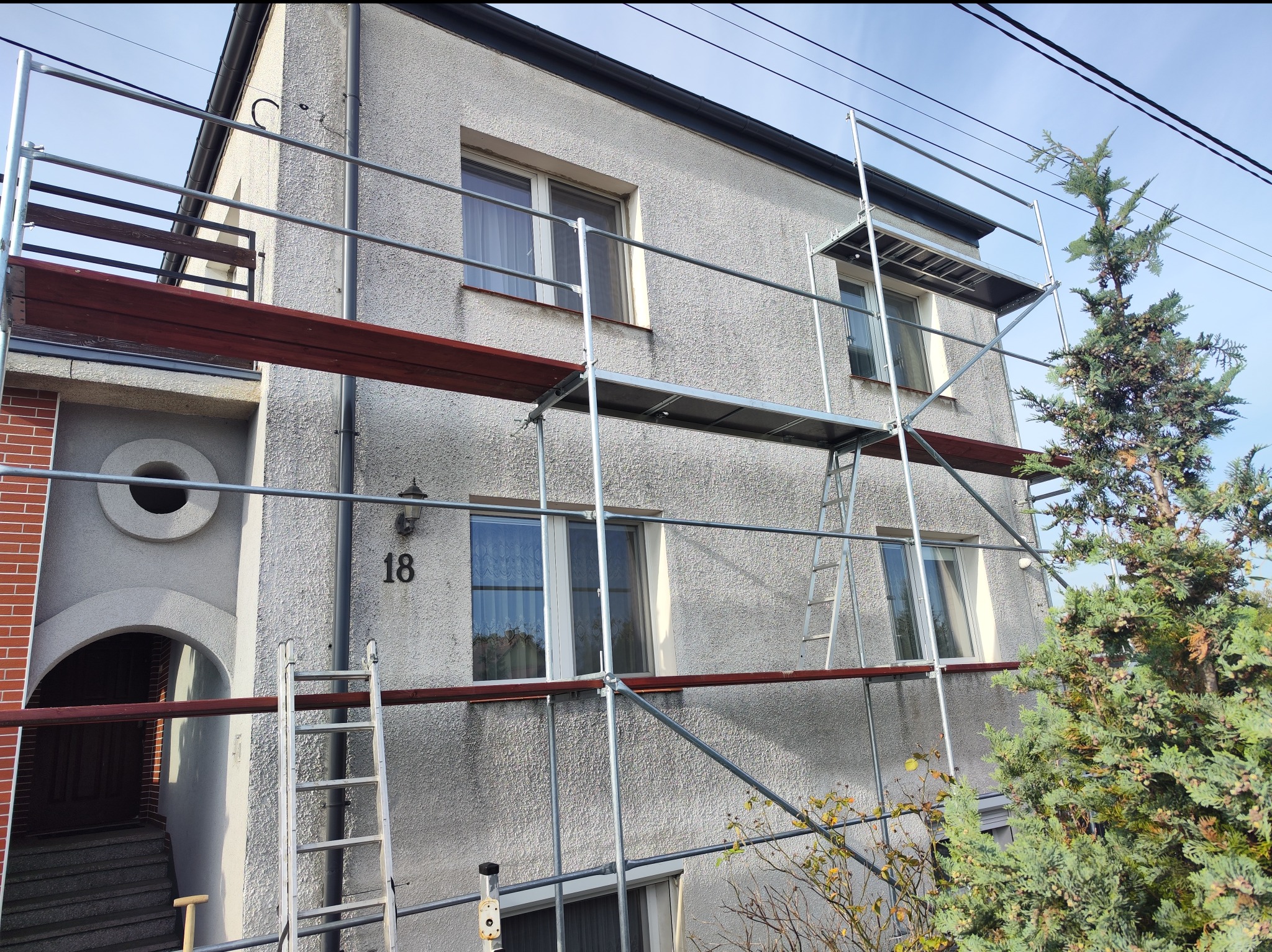 Dwupiętrowy dom z szarą elewacją w trakcie remontu, otoczony rusztowaniem, z widocznym numerem domu '18' i fragmentem zielonego drzewa po prawej stronie.