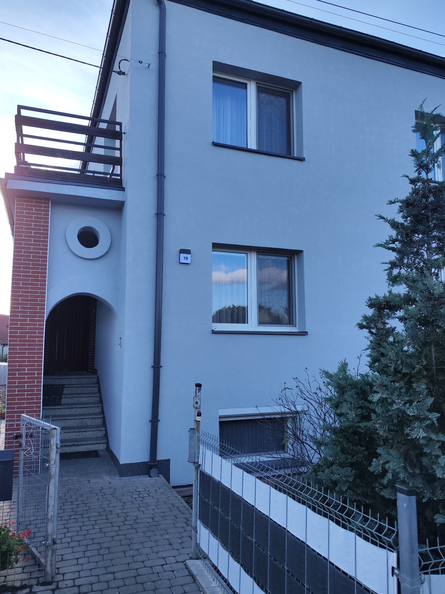 Dwukondygnacyjny dom z elewacją w odcieniach szarości, z balkonem i wejściem z ceglanym elementem, numer domu '18' widoczny na ścianie, obok rośnie iglak.