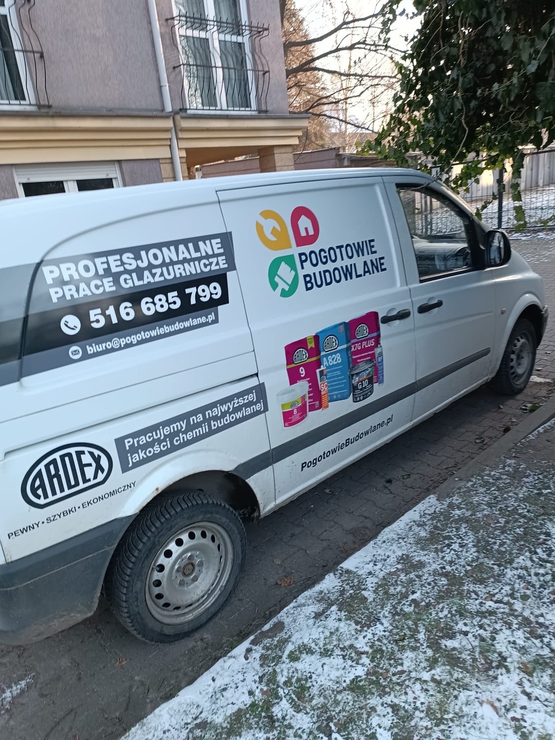 Biały samochód dostawczy z reklamą usług budowlanych, w tym prac glazurniczych, zaparkowany na ulicy z resztkami śniegu. Na karoserii widoczne logo firmy oraz informacje kontaktowe.