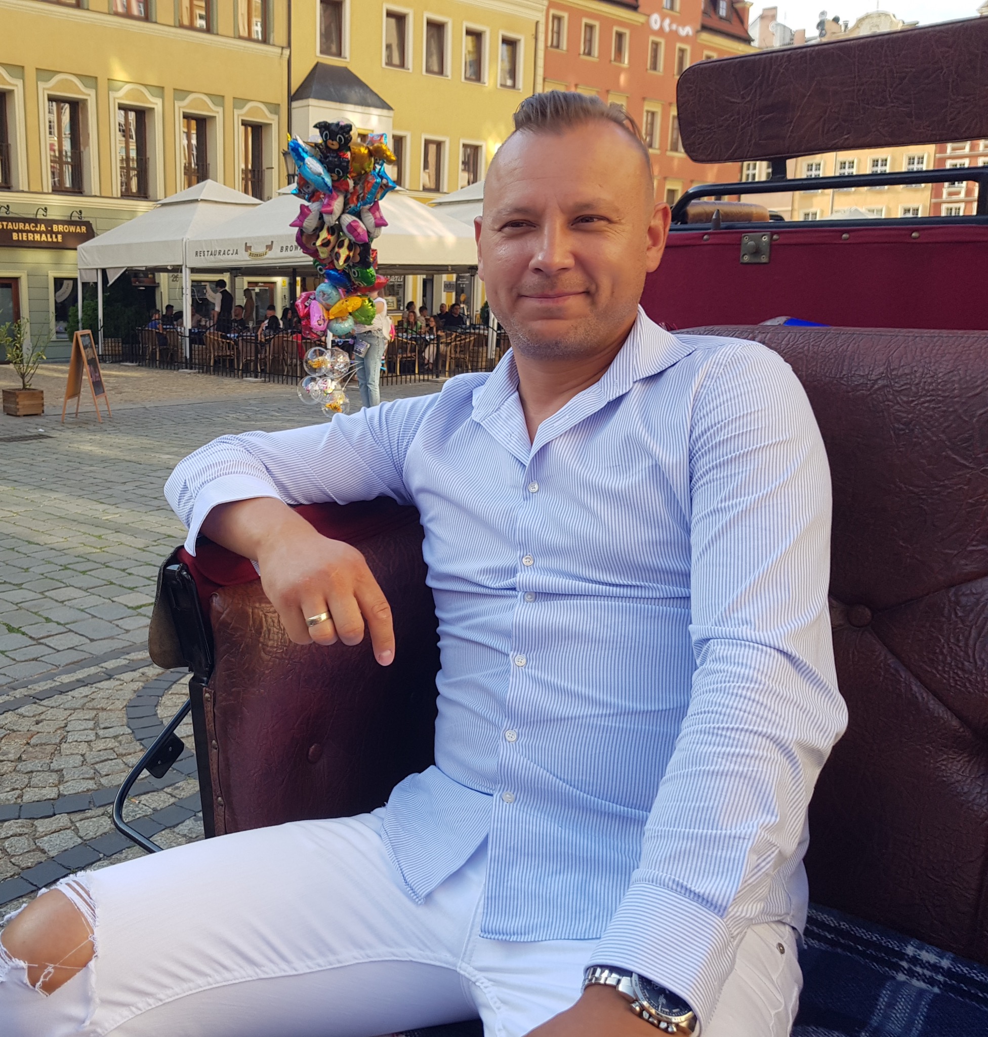 Mężczyzna w niebieskiej koszuli w paski i białych spodniach siedzi w skórzanej karocy na tle miejskiego placu z restauracją i sprzedawcą balonów.