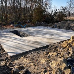 Igor Talarek - Świeżo wylana betonowa płyta fundamentowa z wycięciem na studzienkę, otoczona wykopaną ziemią i niebieskimi rurami, w tle drzewa bez liści w jesiennym słońcu.