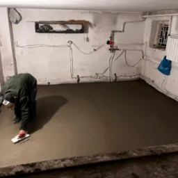 Pracownik w ciemnym pomieszczeniu wyrównuje świeżo wylaną, szarą posadzkę za pomocą pacy, widoczne instalacje na ścianach i okno z kratą.