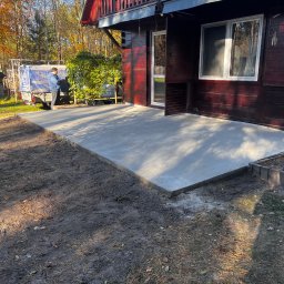 Igor Talarek - Świeżo wylany betonowy taras przylegający do drewnianego domu, widoczny fragment ogrodu i przyczepa z plandeką w tle.