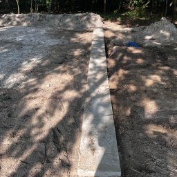 Igor Talarek - Przygotowany wykop pod fundament z ułożonym betonowym elementem szalunkowym i niebieską rurą w tle, widoczne ślady opon.