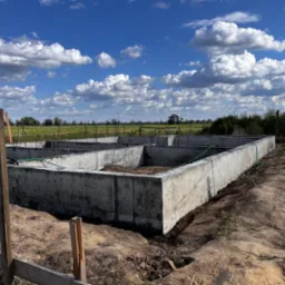 Betonowe ściany fundamentowe na etapie budowy, widoczne z zewnątrz, z zielonym wężem ogrodowym wewnątrz, na tle błękitnego nieba z białymi chmurami i zielonego pola.